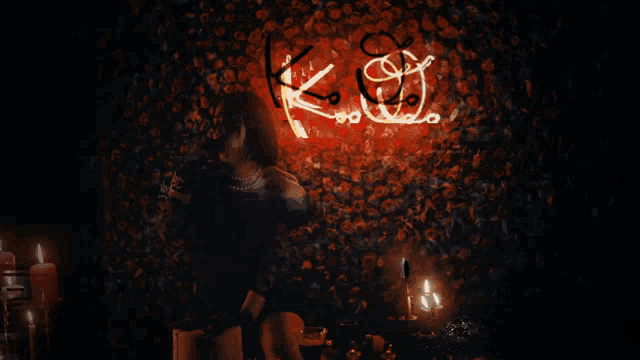 a woman in a black dress drinks from a glass in front of a neon sign that says k j
