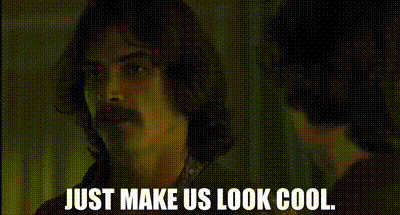 a man with long hair and a mustache is looking at the camera and says just make us look cool .