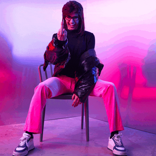 a woman wearing pink pants and a black jacket is sitting in a chair and giving the middle finger