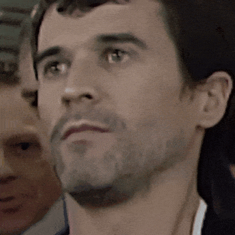 a man with a beard looks at the camera with another man behind him