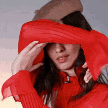 a woman is covering her face with a red scarf while wearing a hat .
