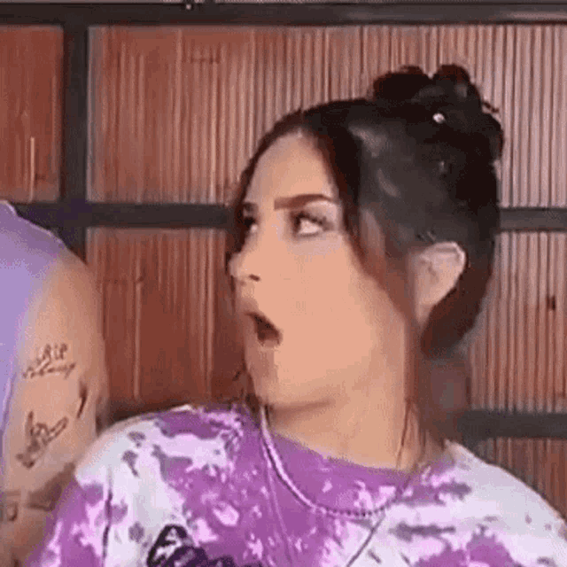 a woman wearing a purple tie dye shirt and a necklace is making a surprised face .