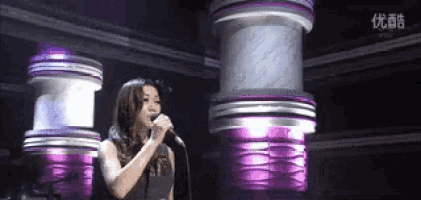 a woman singing into a microphone in front of purple cylinders with chinese writing on them