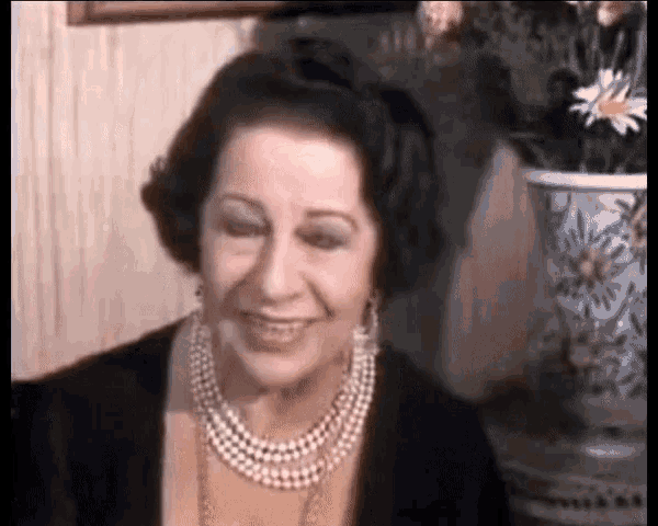 an elderly woman wearing a pearl necklace and earrings is smiling in front of a vase of flowers .