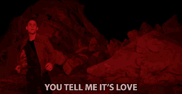 a man in a red suit stands in front of a pile of rocks with the words you tell me it 's love below him