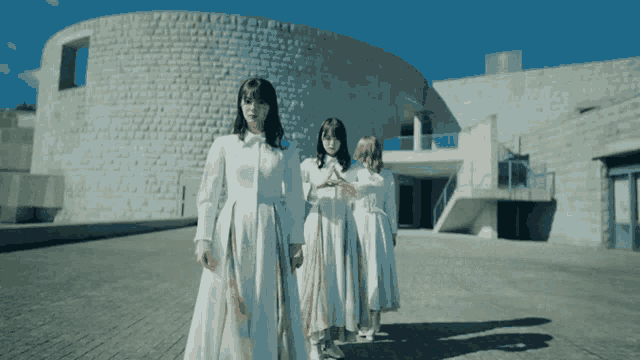 three women in white dresses are walking in front of a large brick building