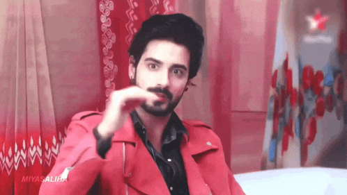 a man with a beard is wearing a red jacket and sitting on a bed .