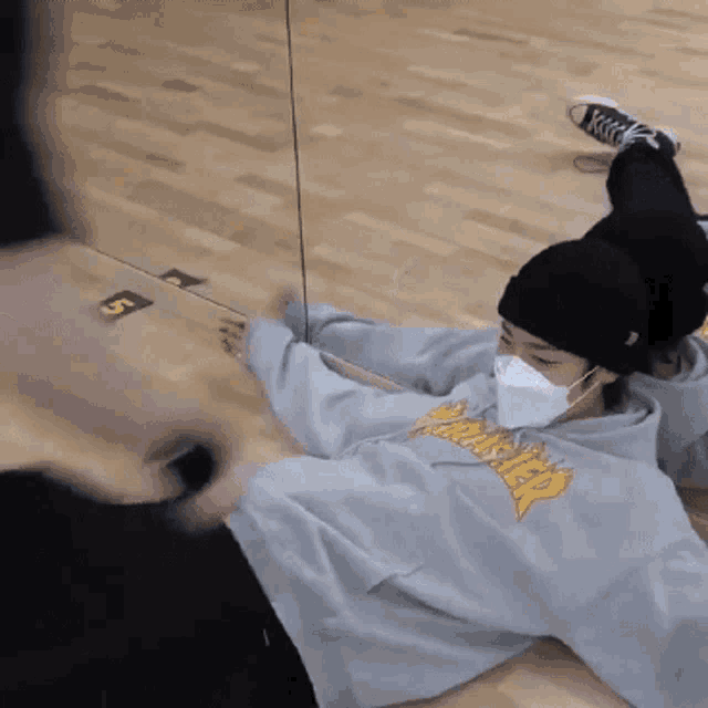 a person wearing a mask and a thrasher sweatshirt is laying on the floor in front of a mirror .
