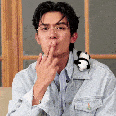 a man wearing glasses and a denim jacket holds his finger to his mouth