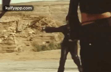 a man is standing next to another man holding a gun in a desert .