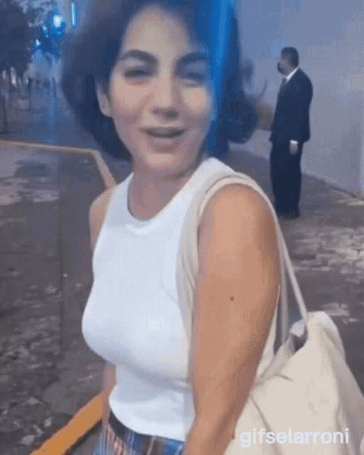a woman in a white tank top is smiling and holding a white purse .