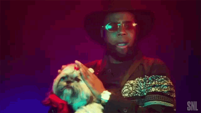 a man wearing sunglasses and a hat holds a small white dog in his arms with the snl logo on his sleeve
