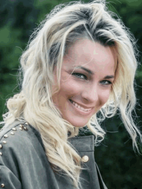 a woman with blonde hair is smiling and wearing a green jacket with studs on the sleeves