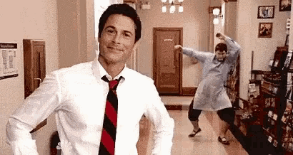 a man in a white shirt and tie is standing next to a woman in a white dress dancing in a hallway .