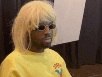 a man wearing a wig and sunglasses is sitting in front of a black curtain .