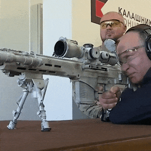 two men are aiming a sniper rifle in front of a sign that says kalash on it
