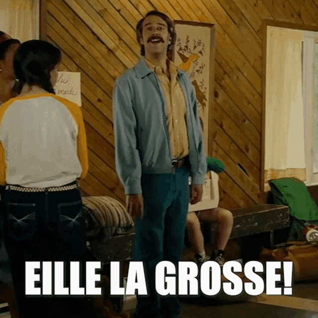 a man with a mustache is standing in a room with the words elle la grosse written on the floor