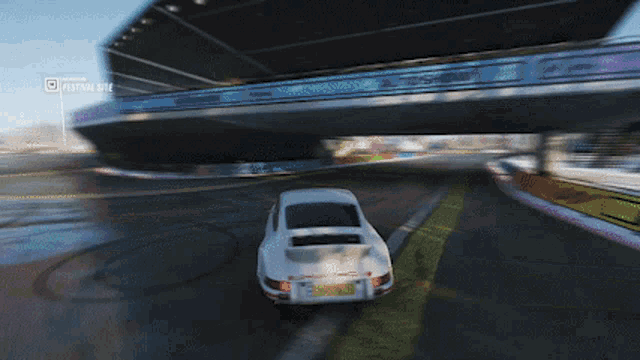 a white car is driving on a wet road under a bridge with a festival site sign above it