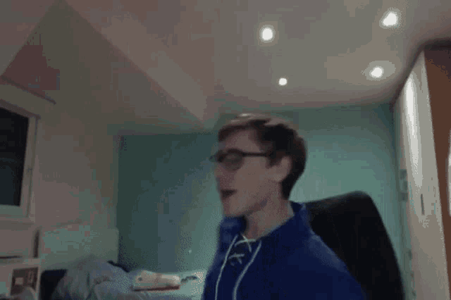 a man in a blue shirt and glasses is standing in a room with a bed .
