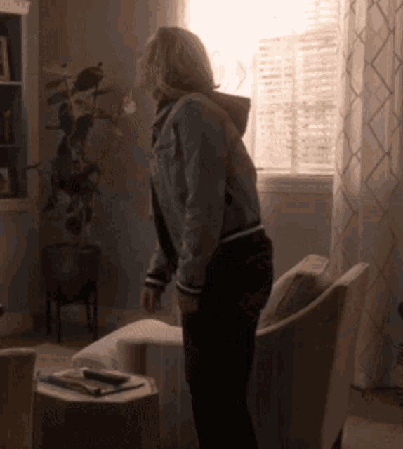 a woman in a denim jacket stands in a living room in front of a window
