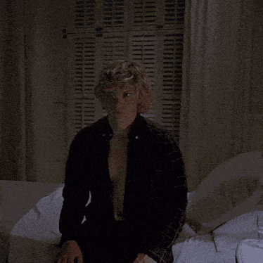 a man in a black shirt sits on a bed in front of shutters
