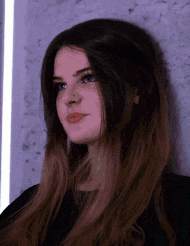 a woman with long dark hair and blue eyes is leaning against a purple wall