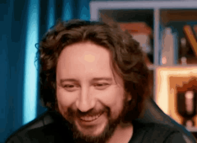 a man with long hair and a beard is smiling in front of a bookshelf .