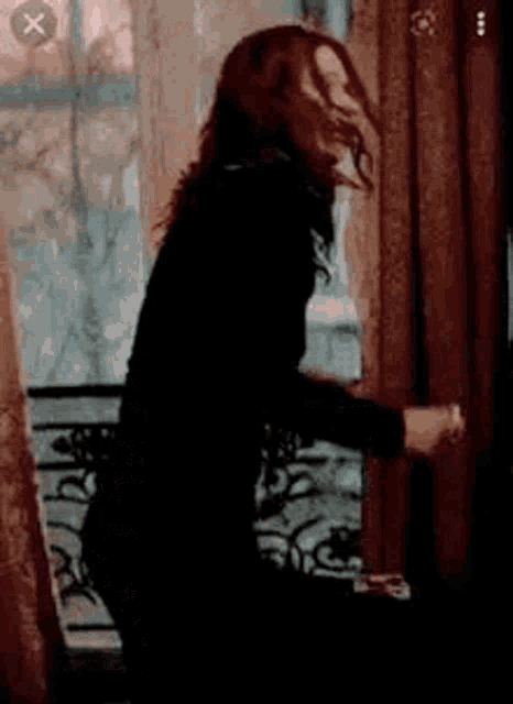 a woman in a black jacket is standing in front of a window holding a curtain .