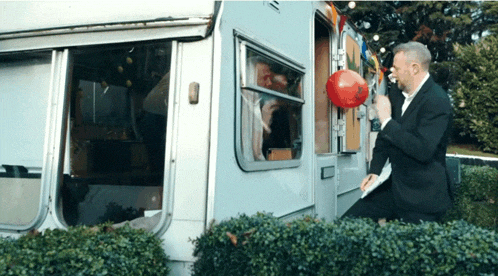 a man in a suit holds a red balloon in front of a camper