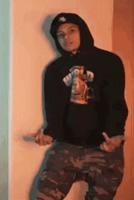 a man wearing a black hoodie and camo pants is standing in front of a wall .