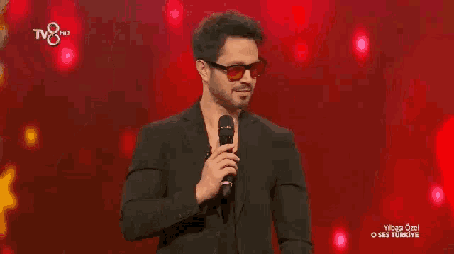 a man in a suit and sunglasses holds a microphone in front of a red background that says tv8 hd on it