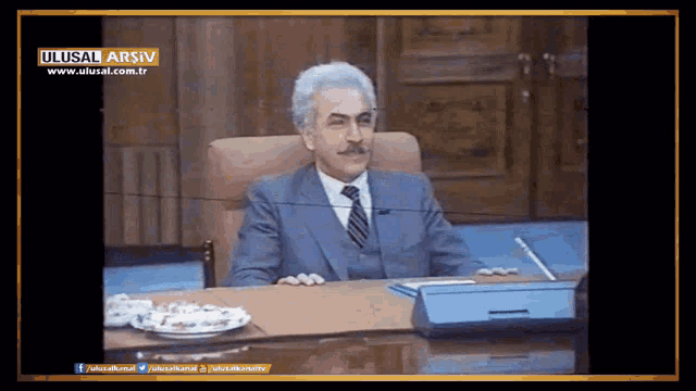 a man in a suit and tie sits at a desk in front of a screen that says ulusal arsiv
