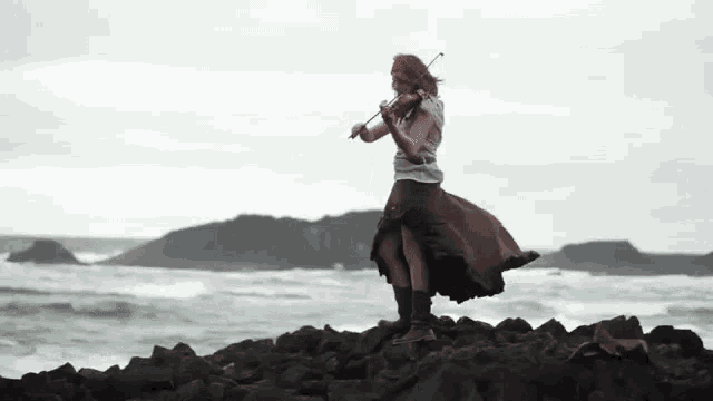 a woman is playing a violin on a rocky shoreline near the ocean