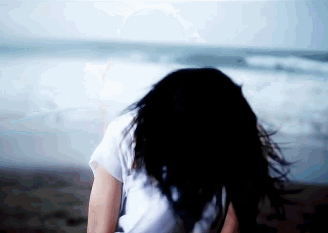 a woman with long black hair is sitting on the beach