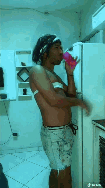 a shirtless man drinking from a pink cup in front of a refrigerator ..