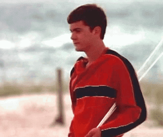 a young man in a red sweater is holding a book while walking on the beach .