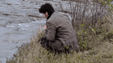 a man in a plaid jacket sits on a grassy shoreline near a river