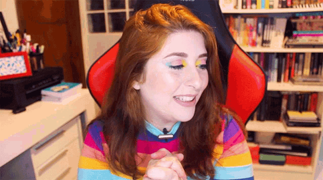 a woman wearing a rainbow striped shirt is sitting in a red chair in front of a bookshelf