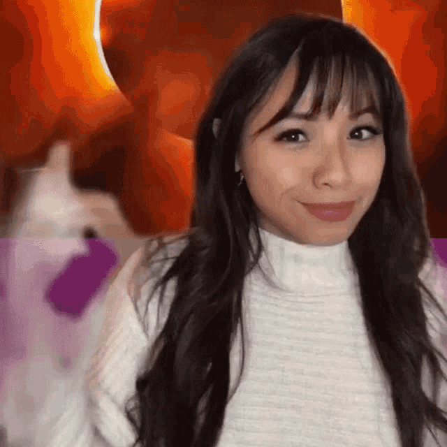 a woman with long hair and bangs is wearing a white sweater and giving a thumbs up .