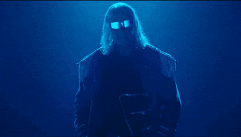a man with long hair and sunglasses is standing in a dark blue room