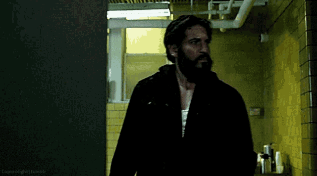 a man with a beard and a black jacket is standing in a hallway .