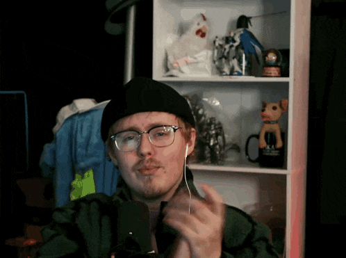 a man wearing glasses and earbuds applauds in front of a shelf with stuffed animals on it