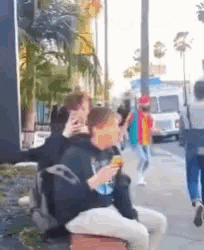 a group of people are walking down a sidewalk while a man sits on a bench eating a sandwich .