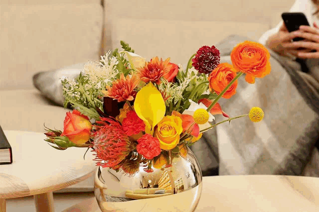 a vase filled with flowers sits on a table next to a person using a cell phone