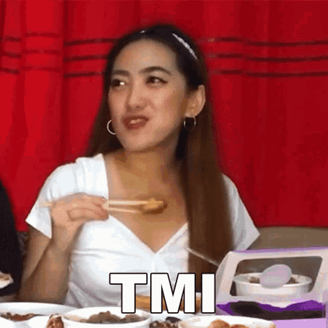 a woman is sitting at a table eating food with chopsticks and the word tmi is on the bottom of the picture .