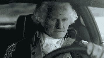 a man dressed as george washington is driving a car with a serious look on his face .