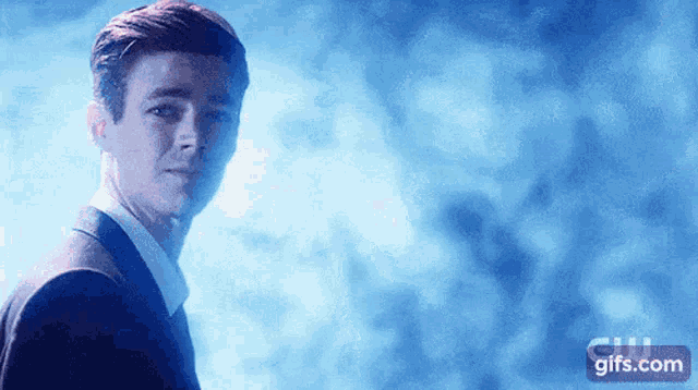 a man in a suit is standing in front of a blue background with a gifs.com logo in the corner