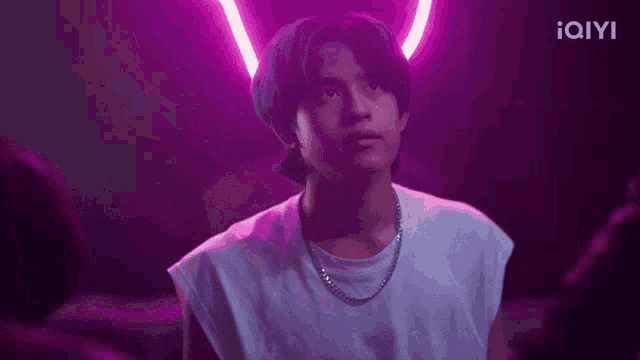 a young man wearing a white tank top and a chain around his neck is standing in front of a purple neon sign .