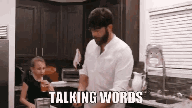 a man is standing in a kitchen talking to a little girl who is drinking a drink .