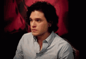a man with long hair is wearing a blue shirt and sitting in front of a red background .